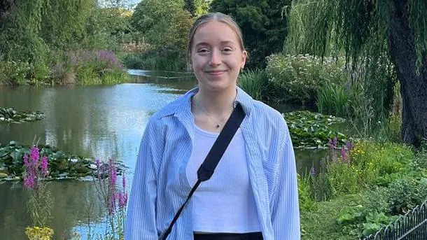 Libby sorrindo para a câmera em frente a um lago com nenúfares, plantas e flores. Ela tem cabelo loiro amarrado para trás e veste uma camisa listrada azul e branca com uma regata branca por baixo. Ela também está usando uma bolsa preta e um colar de prata simples com um pequeno pingente.